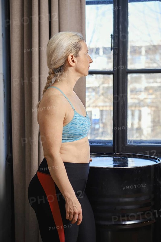 A woman stands near a window, wearing a sports bra and leggings, looking thoughtfully outside. The room is well-lit, highlighting her focused expression as she engages in a morning routine