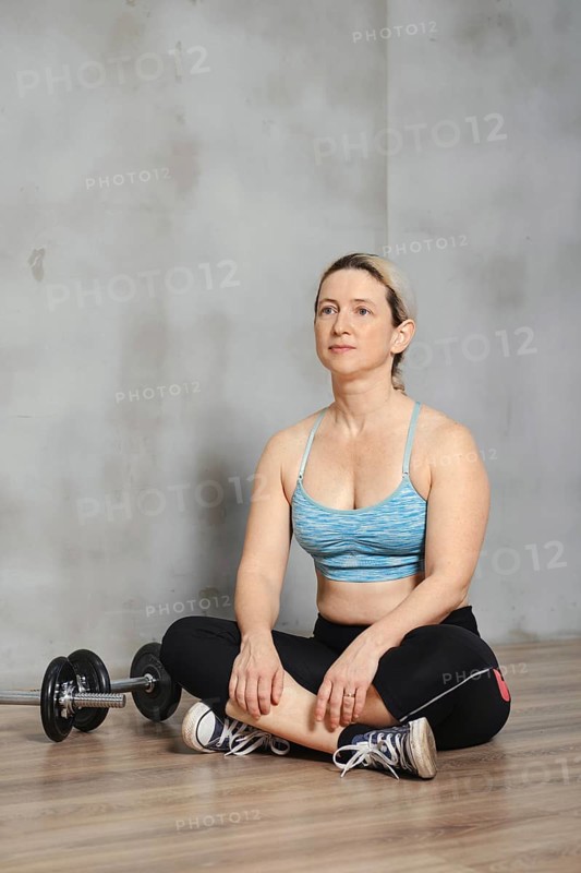 A fit woman is seated cross-legged on the gym floor, looking determined and focused