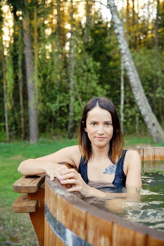 Outdoor portrait of pretty woman in a wooden hot tub surrounded by lush greenery. The warm glow of the evening sun enhances the serene atmosphere, creating a perfect setting for relaxation