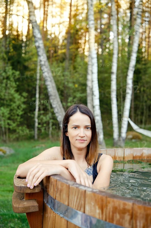 A woman enjoys a tranquil moment in a wooden hot tub, framed by tall trees and lush greenery during sunset. The warm light adds a calming ambiance to her peaceful retreat