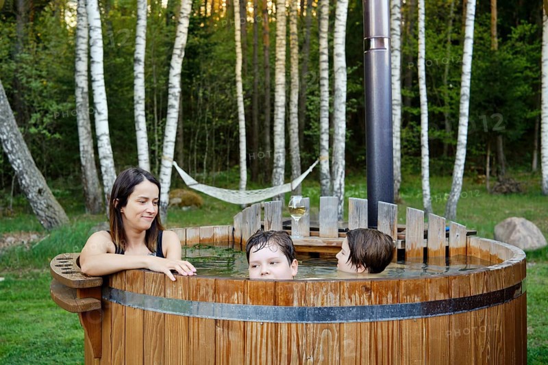 A woman and two children share a joyful moment in a wooden hot tub surrounded by trees. The afternoon sun creates a serene atmosphere, while a hammock hangs in the background for lounging