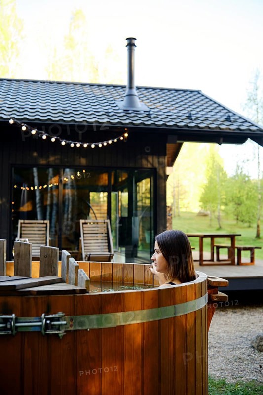 A woman enjoys relaxing in a wooden hot tub in the backyard of a tiny house. The sun illuminates the surrounding greenery, and warm lights add a cozy ambiance to the tranquil setting
