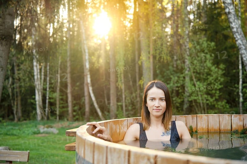A woman enjoys a soothing soak in a wooden hot tub amidst tall trees during a serene sunset. The warm light filters through the leaves, creating a peaceful and calming atmosphere