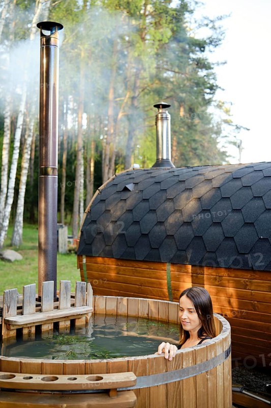 A woman enjoys a peaceful moment in an outdoor wooden hot tub surrounded by tall trees. Steam rises from the sauna, creating a calm atmosphere perfect for relaxation in nature