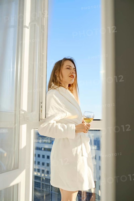 A woman stands on a balcony, dressed in a soft white robe, sipping a glass of beverage. She gazes out at the wide-open sky, basking in the warm sunlight and enjoying a serene moment