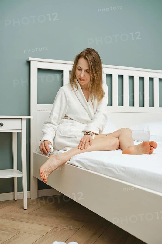 A woman in a white bathrobe peeling dead skin with electric exfoliating machine sitting in bedroom