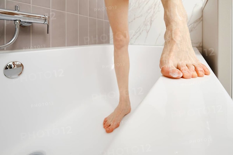 A person balances on the edge of a bathtub, showcasing a moment of daily life. The bathroom features light tiles and a sleek design, emphasizing the simplicity of the setting