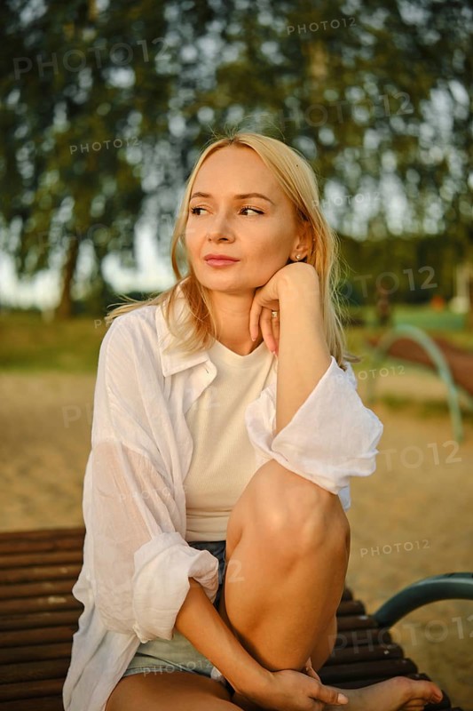 A young woman with blonde hair rests her chin on her hand while sitting on a bench in a park. Sunlight filters through trees, creating a serene atmosphere during the late afternoon hours
