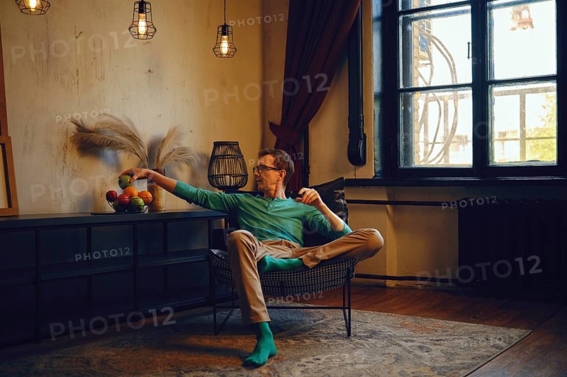 A man sits comfortably in a contemporary chair, casually extending his arm to grab fruit from a nearby table
