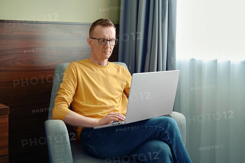 A man sits in a comfortable chair, focused on his laptop in a bright room. Soft natural light streams through the curtains as he engages in his tasks, showcasing a productive atmosphere