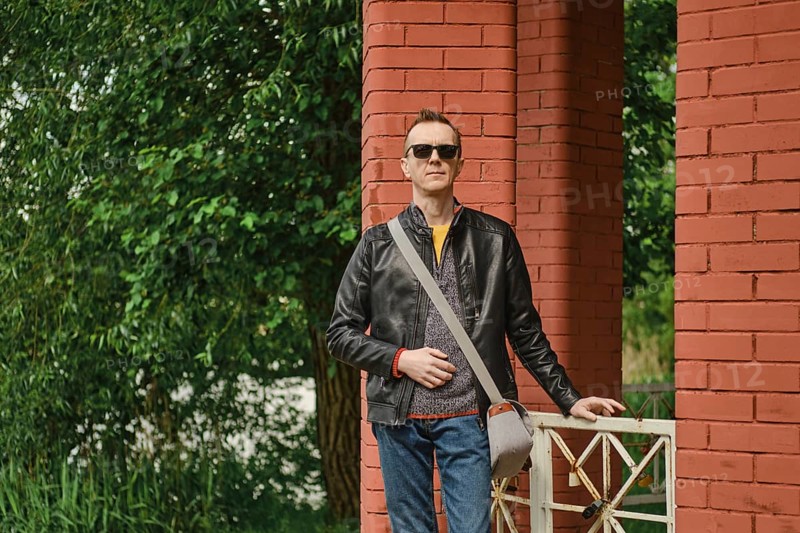 A man stands with a relaxed posture beside vibrant red brick pillars, dressed in a casual outfit featuring sunglasses and a messenger bag