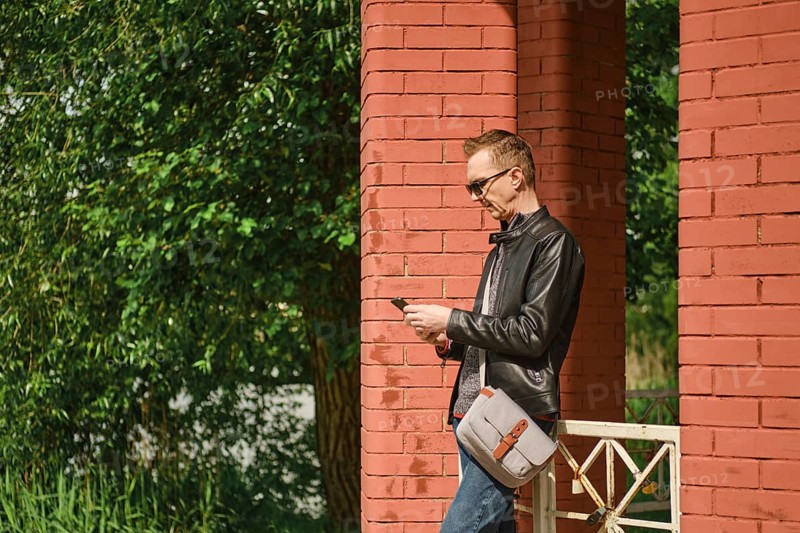 A man wearing sunglasses and a leather jacket stands by red brick columns, attentively using his smartphone. Lush green foliage surrounds the structure, indicating a pleasant sunny day