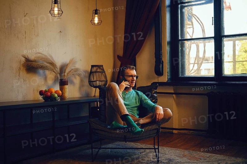 A man dressed in a green long-sleeve shirt and light pants relaxes in a chair, engaging in a phone conversation. The room features warm lighting, a window, and a basket of fruits nearby