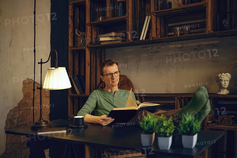 A middle-aged man relaxes with his feet up on a desk in a home office, reading a book. Natural light illuminates the cozy space, filled with books and decorative elements