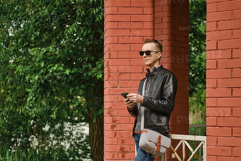 A man dressed in a leather jacket and sunglasses stands by red brick columns. He holds a smartphone and looking for an approaching taxi