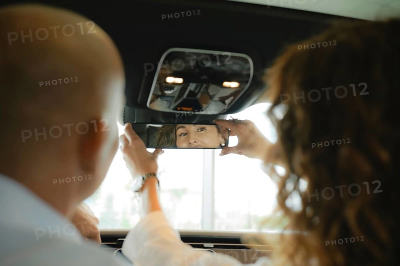 The view from the rear seat of the car is a couple of sitting in front. A woman sets up a rear view mirror and smiles