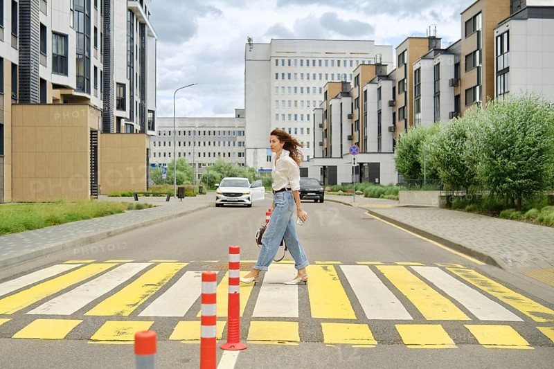 A woman with long hair walks across a marked pedestrian crossing in a contemporary urban area. She wears casual clothing and carries a bag