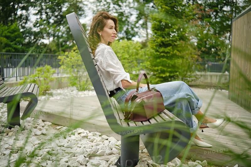 A woman sits comfortably on a bench in a park, surrounded by greenery and decorative stones. She has a stylish handbag and is enjoying the peaceful atmosphere on a bright day