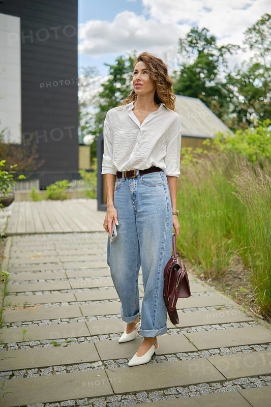 A young woman is walking along a stone path, wearing a white shirt and light blue baggy jeans. She carries a burgundy handbag and checks her phone while surrounded by greenery and modern buildings