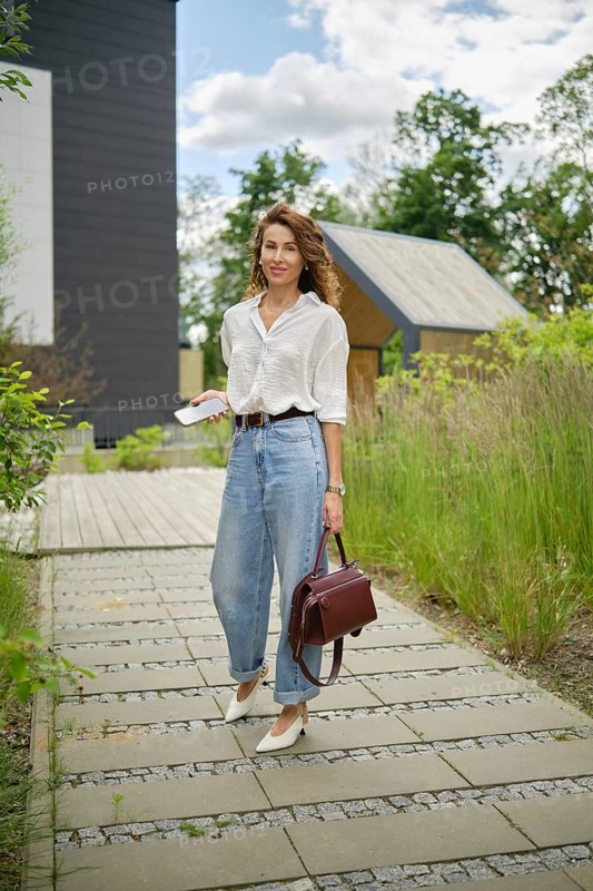 A fashionable woman strolls along a scenic pathway lined with lush plants. She wears a white shirt and high-waisted jeans, carrying a bag and checking her phone while enjoying the outdoors