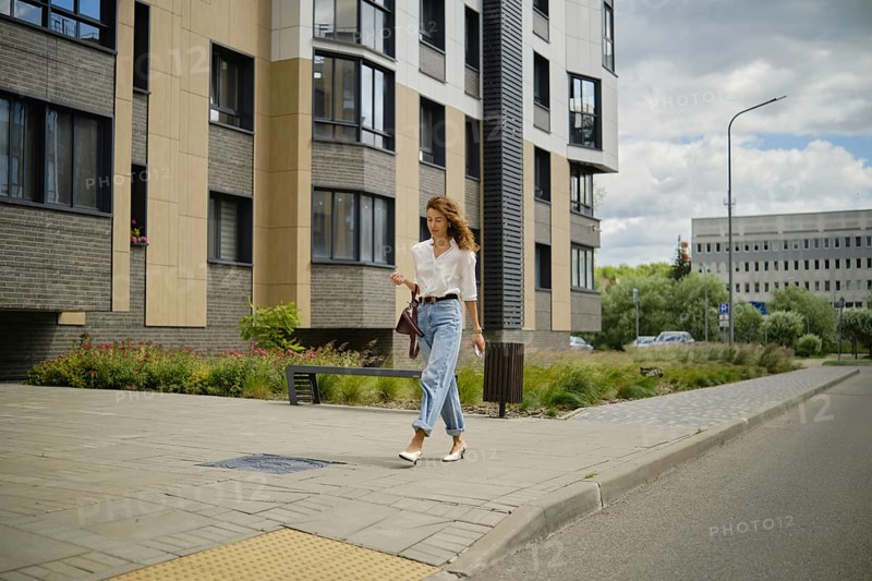A woman strolls down a city sidewalk on a sunny day. She wears a casual outfit with light jeans and a blouse. Modern buildings and lush plants create a vibrant urban atmosphere
