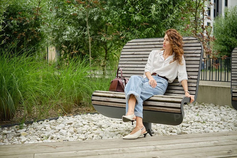 A woman with curly hair is seated on a modern wooden bench in a landscaped urban park. She is wearing a casual outfit, enjoying the surrounding greenery and the warm daylight