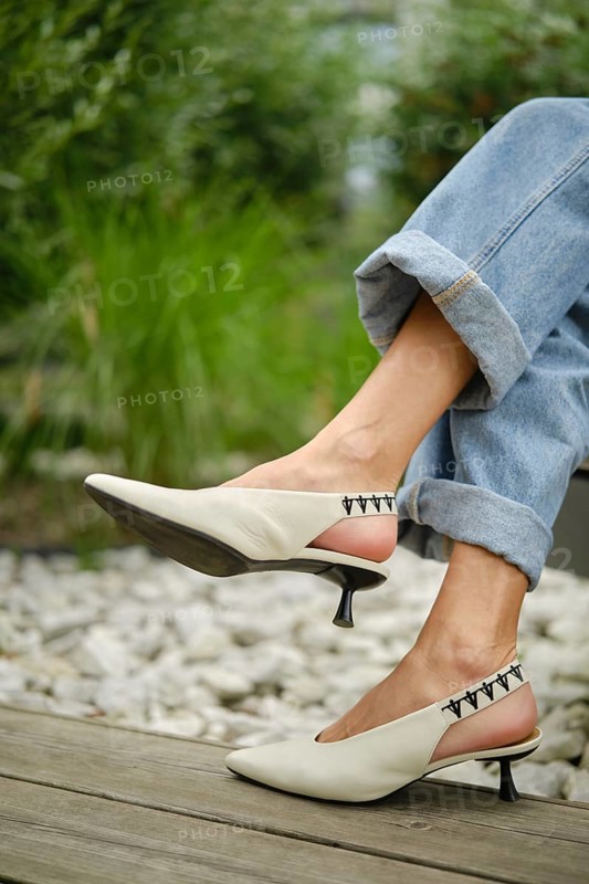 A woman showcases fashionable white slingback heels while sitting outdoors. She wears rolled-up denim jeans, surrounded by lush greenery and pebbles, exuding a casual yet chic vibe