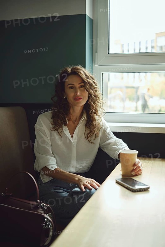 A woman with curly hair sits at a wooden table in a cafe, holding a paper cup. Sunlight streams through the window, illuminating her casual outfit as she seems relaxed and content