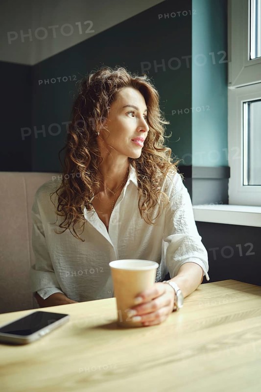 A woman with curly hair sits at a wooden table in a cozy cafe, holding a coffee cup. She gazes out the window, lost in thought, as natural light brightens her surroundings