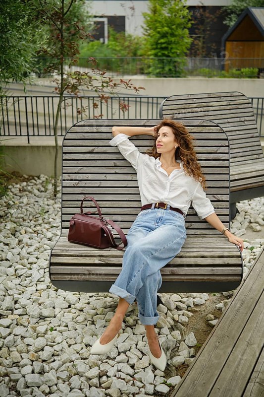 A woman enjoys her time on a stylish bench in a contemporary outdoor area filled with greenery. She wears a white shirt and high-waisted jeans, looking relaxed while holding a handbag
