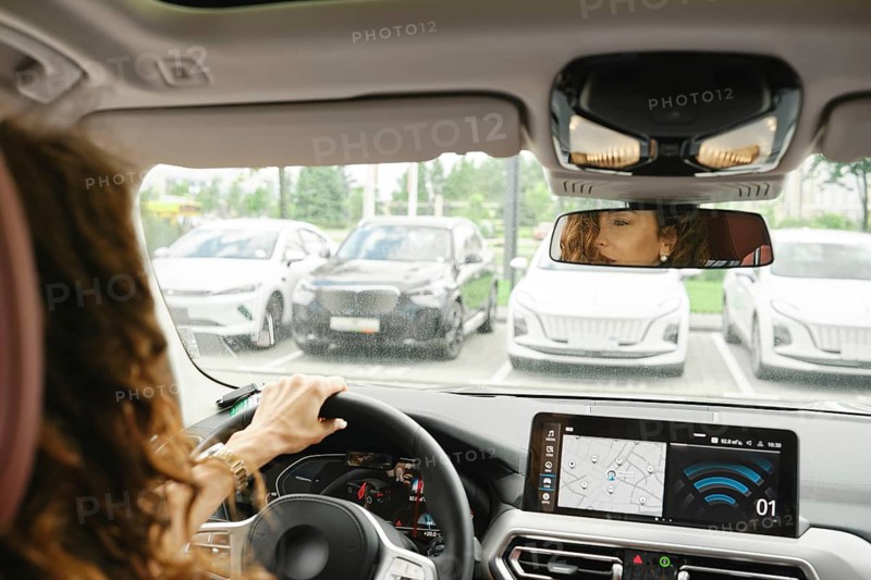 A woman is driving her car in a parking lot, attentively looking at her surroundings through the rearview mirror. A navigation system displays directions on the dashboard, aiding her in parking, AI generated