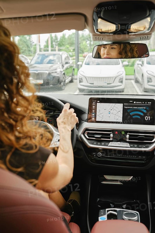 A woman with curly hair is focused on driving a car, checking the navigation screen. She is inside a modern vehicle parked among several other cars on a clear day, AI generated