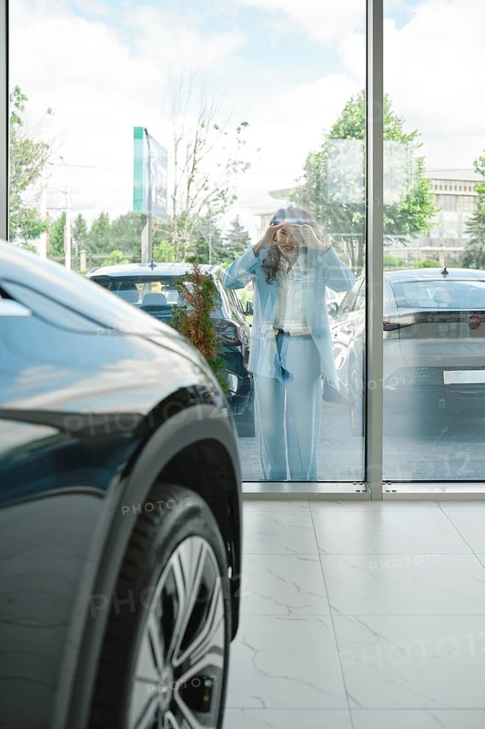 A woman stands outside looking in through large glass windows to a car dealership, admiring various cars displayed. The atmosphere is bright and inviting with a clear sky above, AI generated