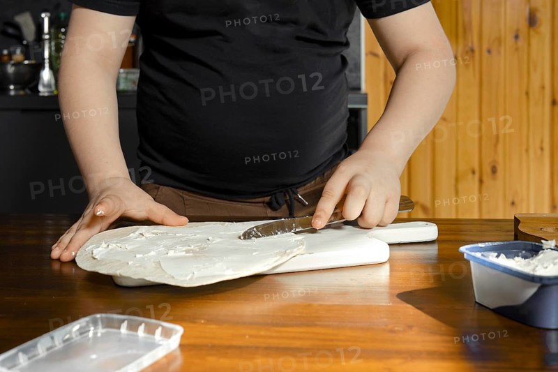 A young boy is spreading a cream cheese onto a flatbread in a well-lit sunny kitchen. He is making simple and healthy snack