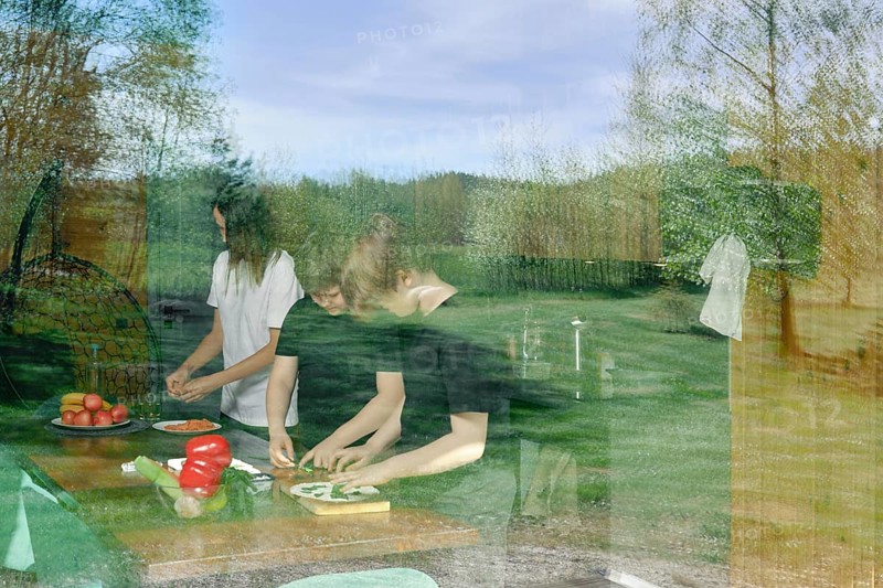 The view through the window to the kitchen, where the family is preparing a light snack. The glass reflects the meadow and the forest where the country house is located