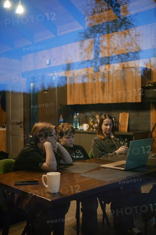 A cozy setting captures two children and an adult focused on a laptop screen inside a modern home. The warm wood interior reflects a comfortable atmosphere during the evening