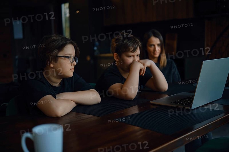 Two children and their mom watching movie on a laptop. They sit at a wooden table in a comfortable indoor environment. Evening light fills the space