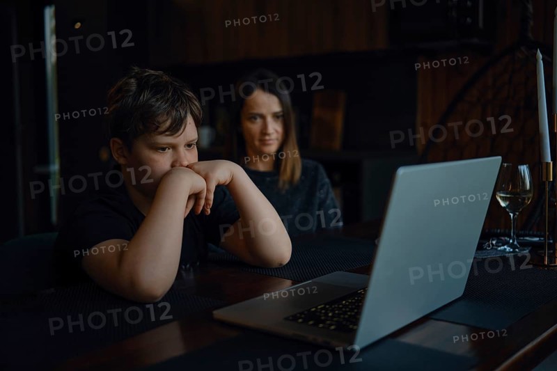 A boy sits focused at a table, watching a laptop screen closely while his mother looks on with a supportive expression