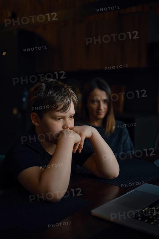 A young boy sits at a table, staring intently at a laptop screen with his hands on his chin. His mother watches him closely from behind, both in a cozy, dimly lit room