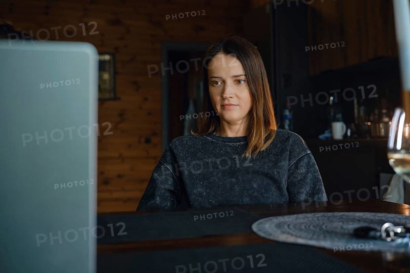 A woman is focused on her laptop while sitting at a wooden table in a warm dark living room. The cozy interior features a rustic design, enhancing her home office atmosphere