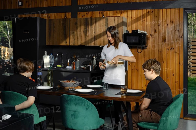 In the kitchen, a woman pours a drink, while two children are sitting at the dining table, impatiently waiting for their meal. The cozy atmosphere demonstrates a family pastime focused on healthy eating