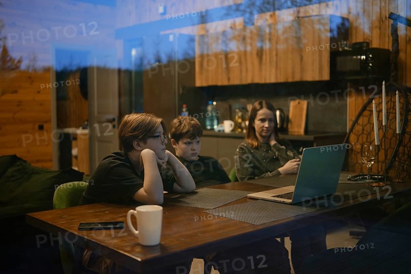 Inside a cozy cabin, a family gathers around a table enjoying a shared experience. Two children are focused on a laptop, while an adult casually checks a phone, creating a warm atmosphere
