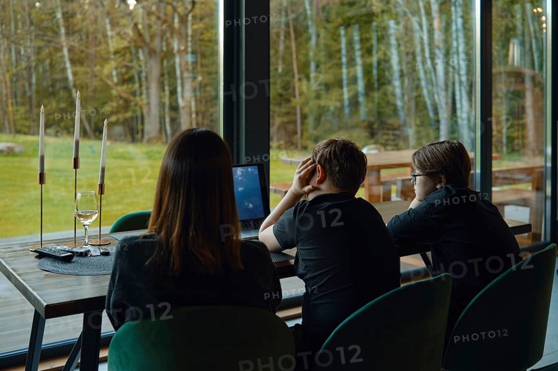 In a cozy lodge surrounded by nature, a family gathers at a table. Two children sit in front of a laptop, focused on their screen. An adult watches them, creating a warm family moment
