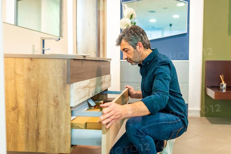 Mature man crouching, opening drawers of a bathroom vanity, checking quality and organization in a home improvement store showroom