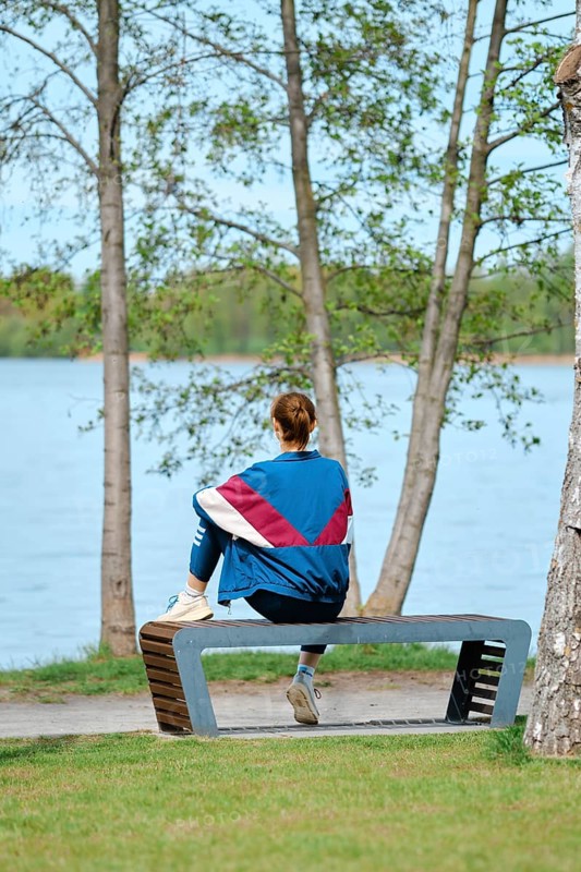 A woman sits on a unique bench facing a serene lake, surrounded by trees. She enjoys the tranquil setting, soaking up the calm atmosphere during a sunny day. Nature's beauty enhances the moment
