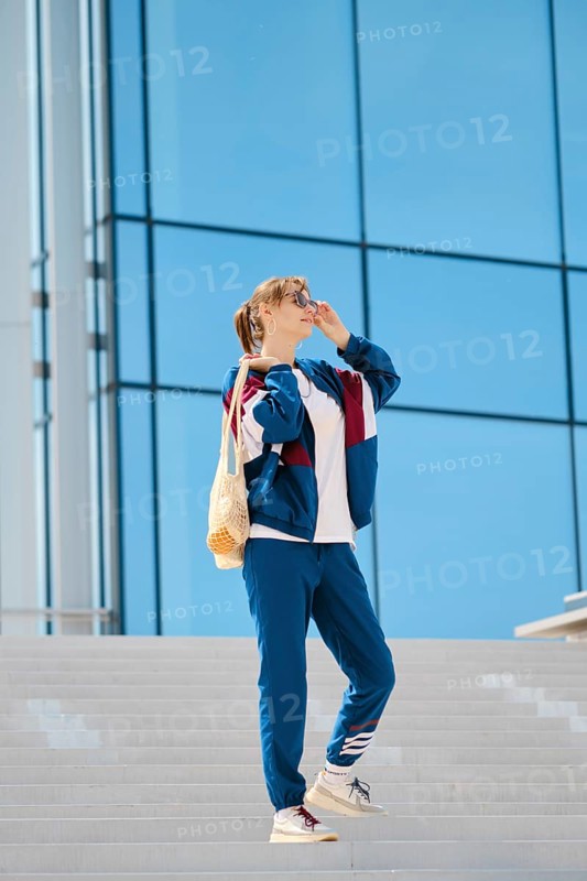 A young individual stands on a staircase, wearing sporty clothing and sunglasses. The sunny weather complements the vibrant blue building behind, creating a lively urban atmosphere