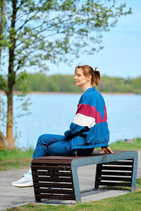 A young woman relaxes on a modern bench near a serene lake, dressed in colorful activewear. The bright sun and blue sky enhance the tranquil atmosphere, highlighting her joy in nature
