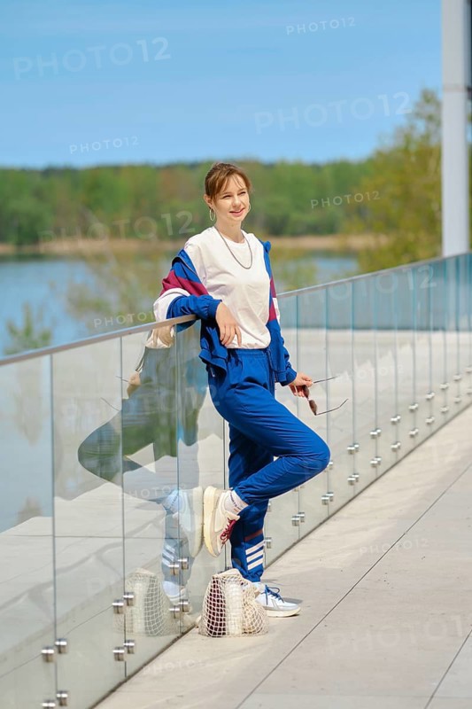A woman dressed in comfortable sportswear leans casually against a glass railing overlooking a serene body of water