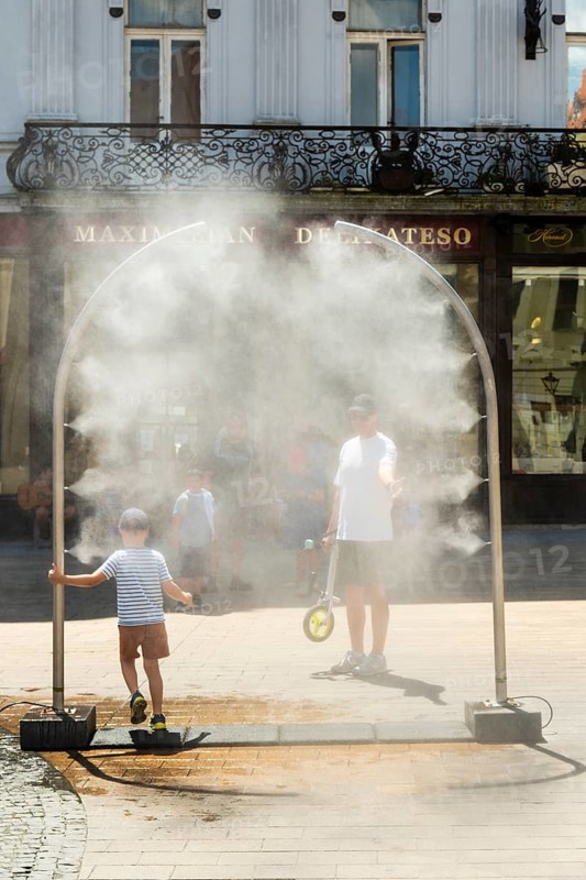 A mobile spray arch sprays a fine mist of water on a sunny square. Passers-by, including a child and a man on a scooter, take advantage of the refreshment in the summer heat, Bratislava, Slovakia
