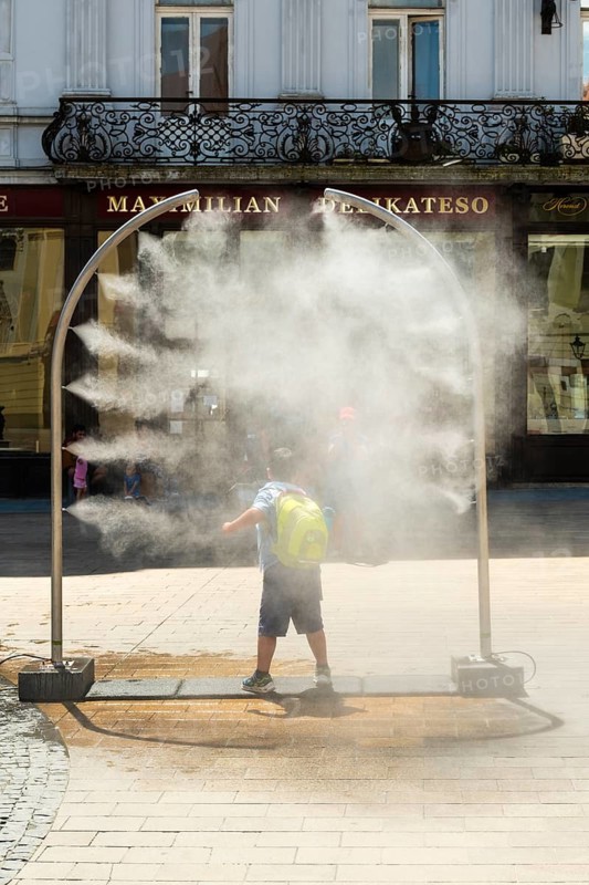 A mobile spray arch sprays a fine mist of water in a sunny square. A child uses the refreshment in the summer heat, Bratislava, Slovakia